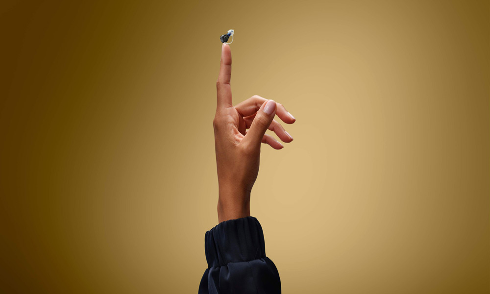 En mycket liten svart Oticon Zeal hörapparat som balanserar på spetsen av ett uppsträckt pekfinger.