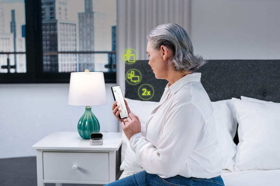 En kvinna med uppsatt grått hår sitter på en säng och tittar på sin smartphone. Hon bär en vit skjorta och har en synlig hörapparat på örat. Runt telefonen visas digitala gröna ikoner som illustrerar trådlös anslutning.