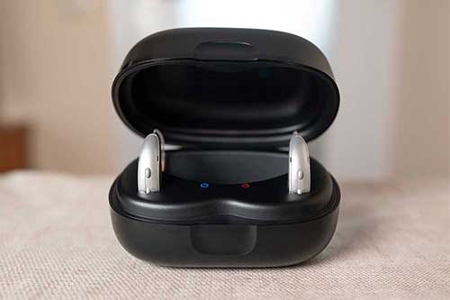 Laddningsbara hörapparater placerade i en svart laddningsstation med öppet lock, stående på ett tygklätt bord i hemmiljö.