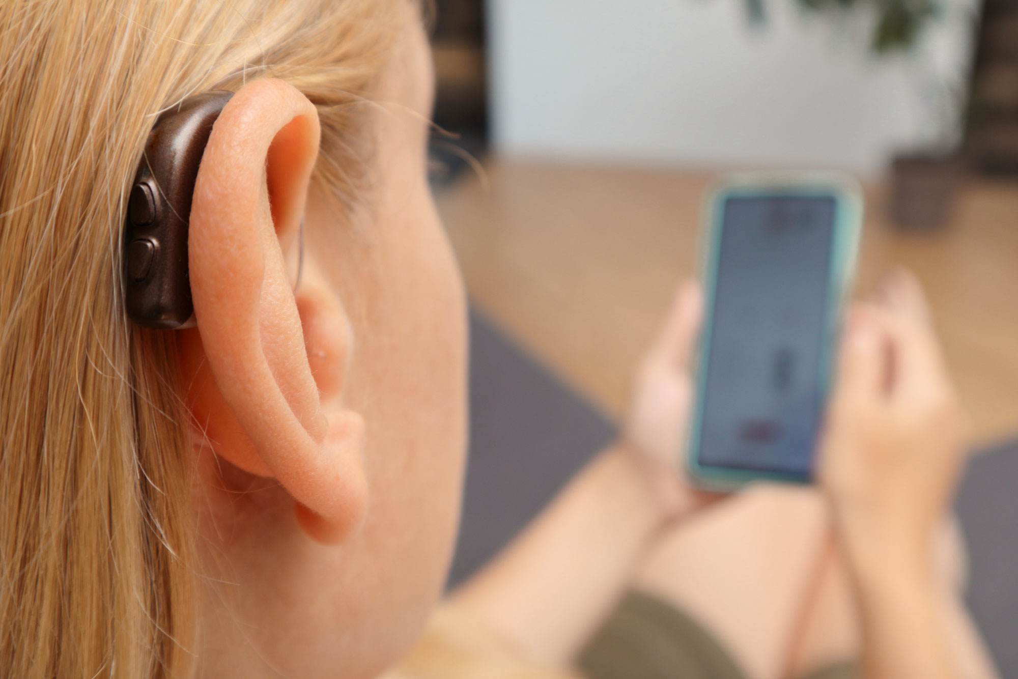En närbild som visar en brun hörapparat placerad bakom örat på en person med ljust hår. I bakgrunden syns personens händer som håller i en smartphone.