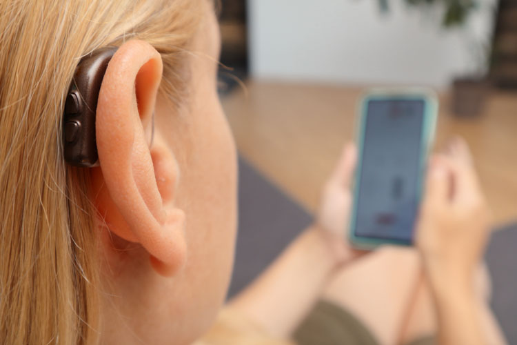 En närbild som visar en brun hörapparat placerad bakom örat på en person med ljust hår. I bakgrunden syns personens händer som håller i en smartphone.