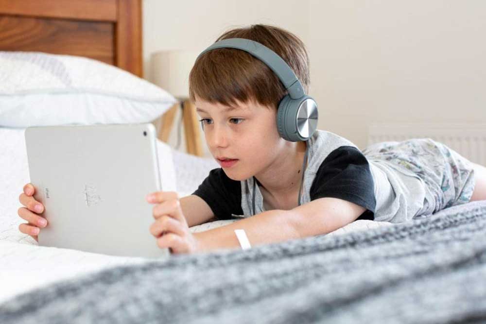 Ett barn i en grå pyjamas med svarta ärmar  ligger på mage i en vitbäddad säng och kollar på en iPad. Han har grön/blåa over-ear hörlurar och brunt hår.