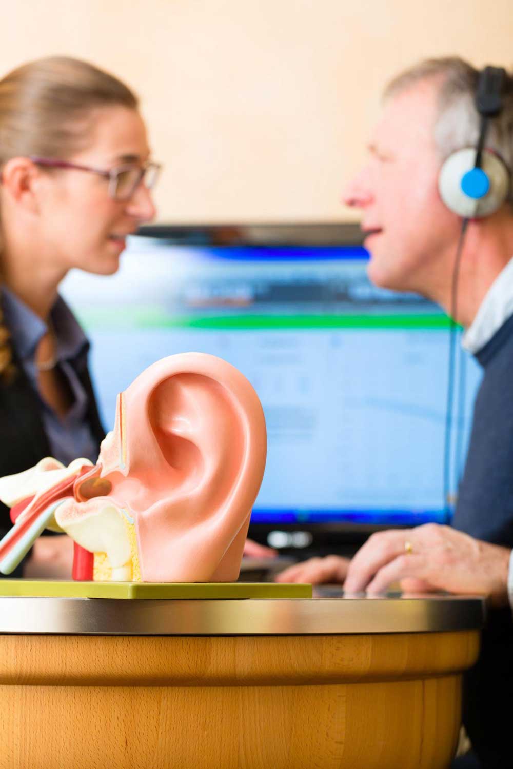 En äldre man genomgår ett hörseltest med hörlurar på, medan en audiolog sitter mittemot honom och ger instruktioner. En modell av ett mänskligt öra är i förgrunden, vilket illustrerar örats anatomi.