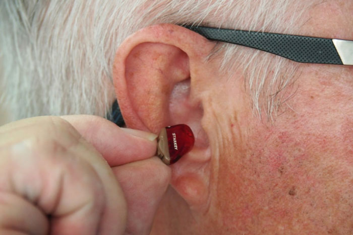 Äldre man med glasögon som sätter in en röd hörapparat i örat.