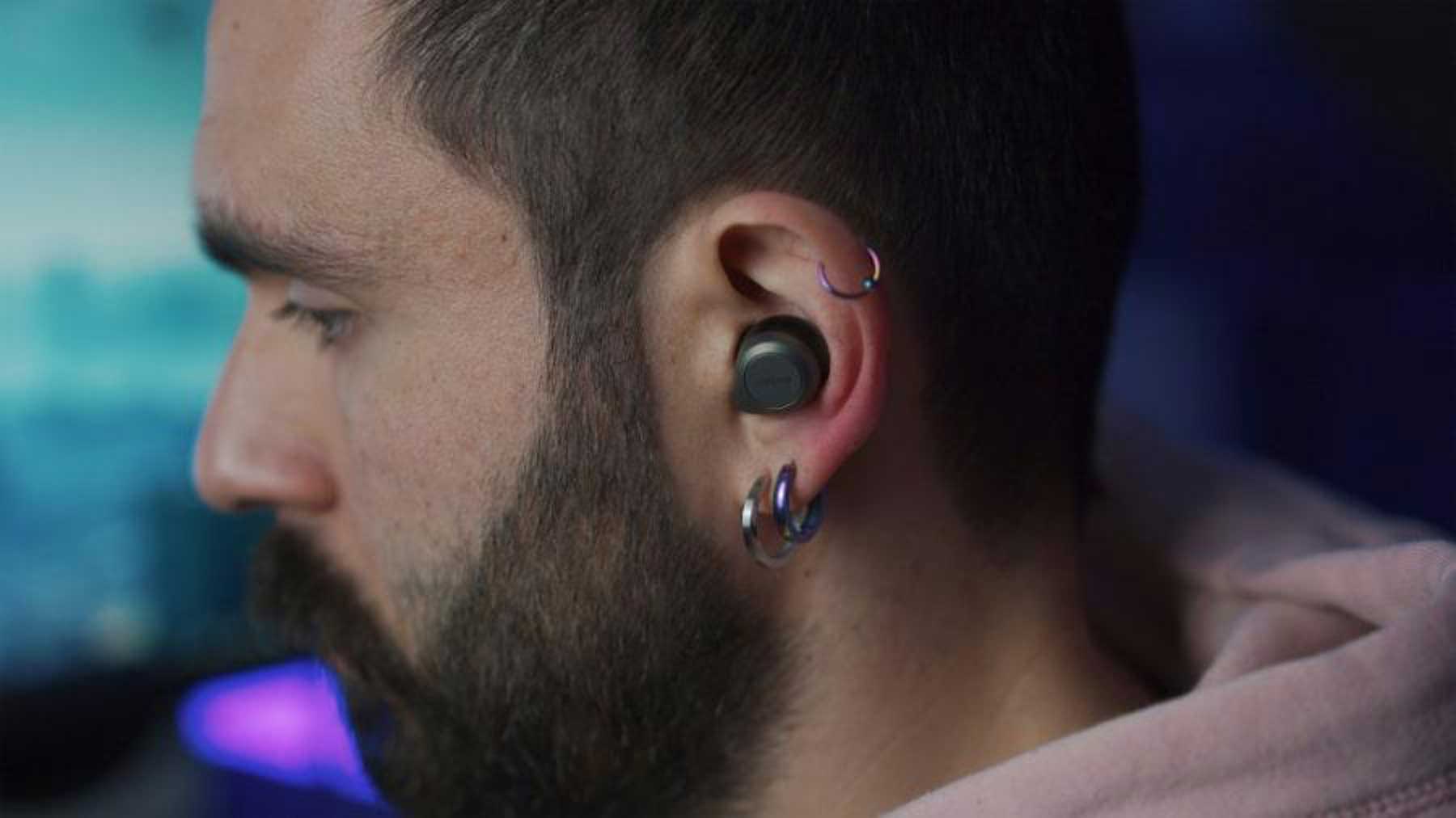 Man med in-ear hörlur och tre metalliska örhängen i örat. Mannen har mörkt kort hår, skägg och en ljusrosa hoodie på sig.