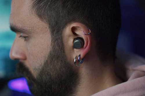 Man med in-ear hörlur och tre metalliska örhängen i örat. Mannen har mörkt kort hår, skägg och en ljusrosa hoodie på sig.